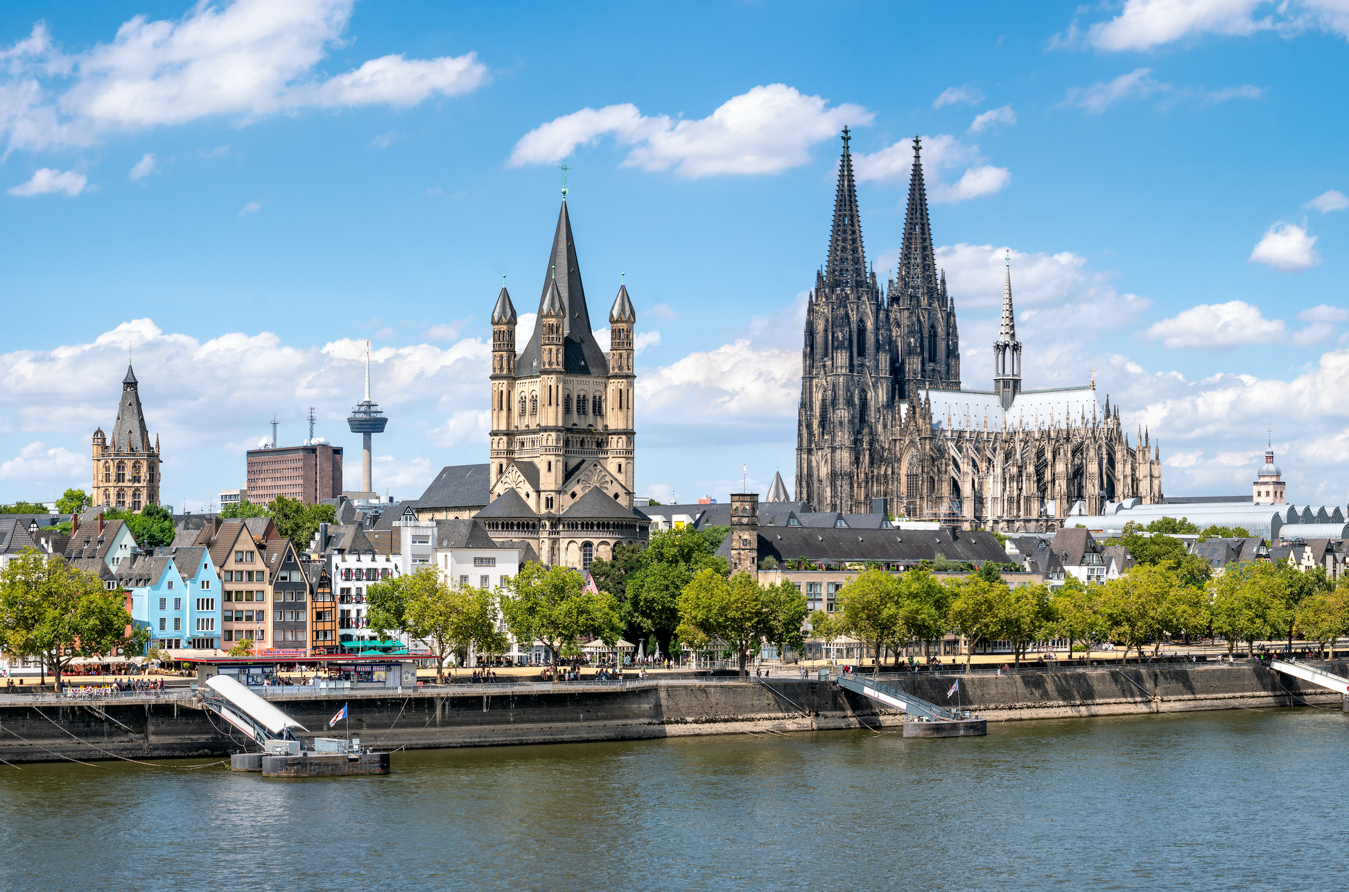 Panoramablick auf das Rheinufer in Köln mit dem Kölner Dom und der Kirche Groß St. Martin, im Hintergrund der Colonius Fernsehturm und andere städtische Gebäude unter einem blauen Himmel mit weißen Wolken.