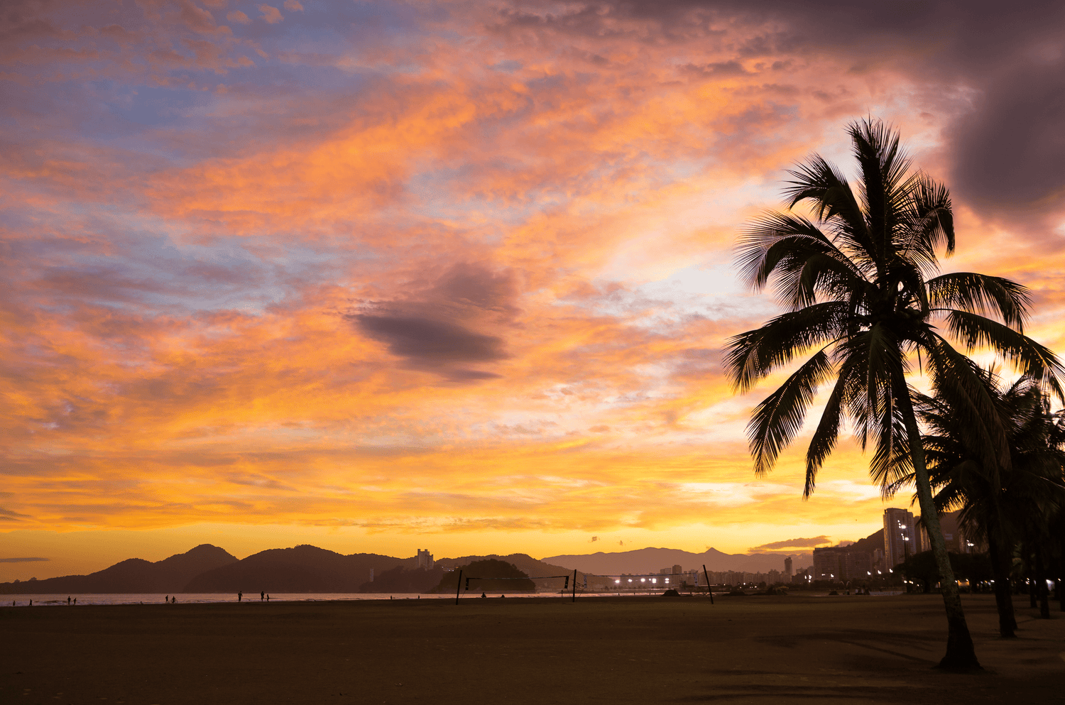 Ein wunderschöner Sonnenuntergang am Strand von Santos, Brasilien. Der Himmel ist in warmen Orangetönen und sanften Rosa- und Blauschattierungen gefärbt. Eine Silhouette einer Palme steht im Vordergrund, während im Hintergrund die Umrisse von Bergen und Gebäuden zu sehen sind. Menschen spazieren am Strand entlang und genießen die friedliche Atmosphäre.