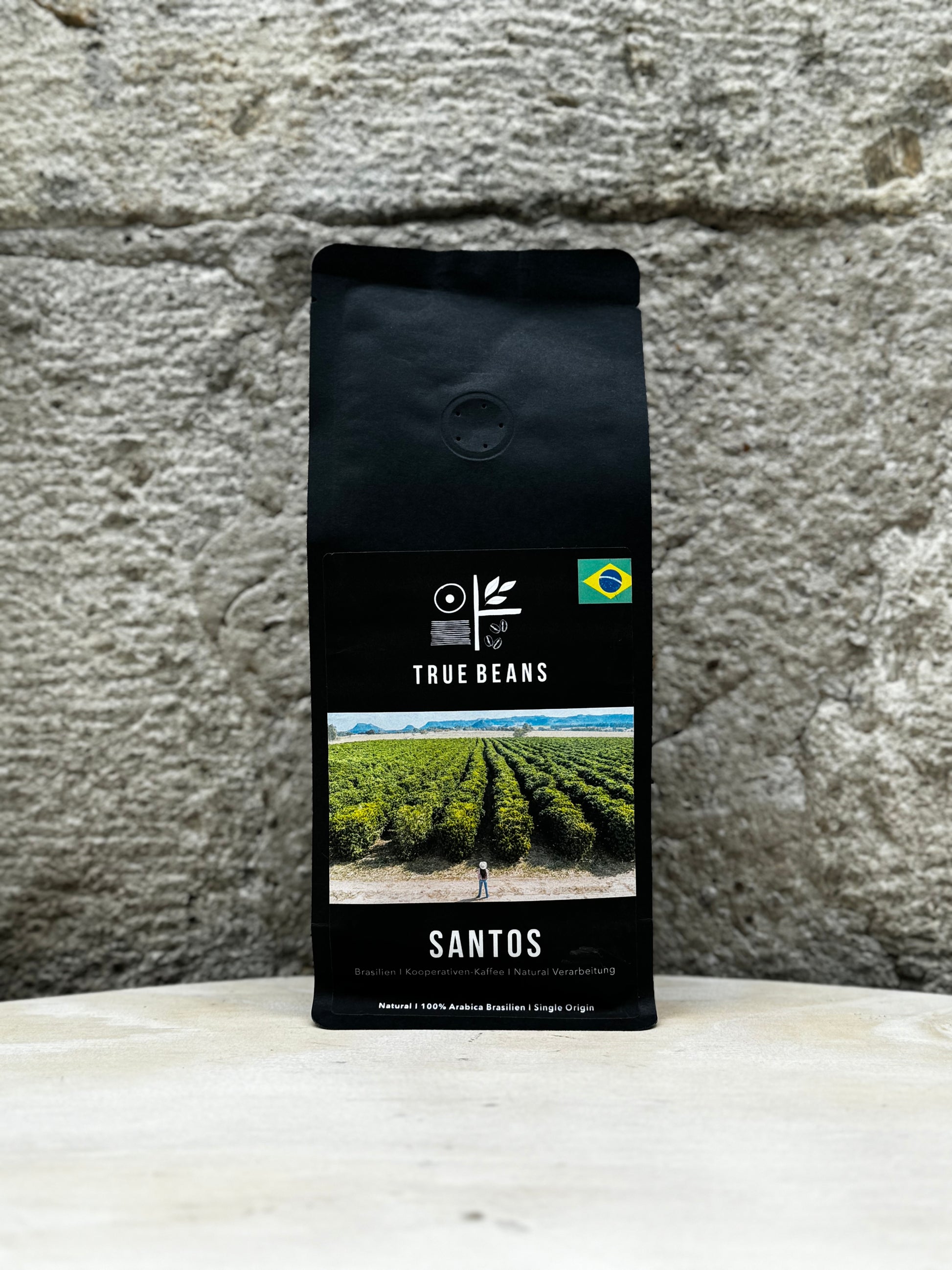 Eine Packung True Beans Santos Kaffee vor einer Steinwand. Auf der Verpackung ist ein Bild einer Kaffeefarm in Brasilien zu sehen, sowie die Beschreibung "Brasilien | Kooperativen-Kaffee | Natural Verarbeitung".