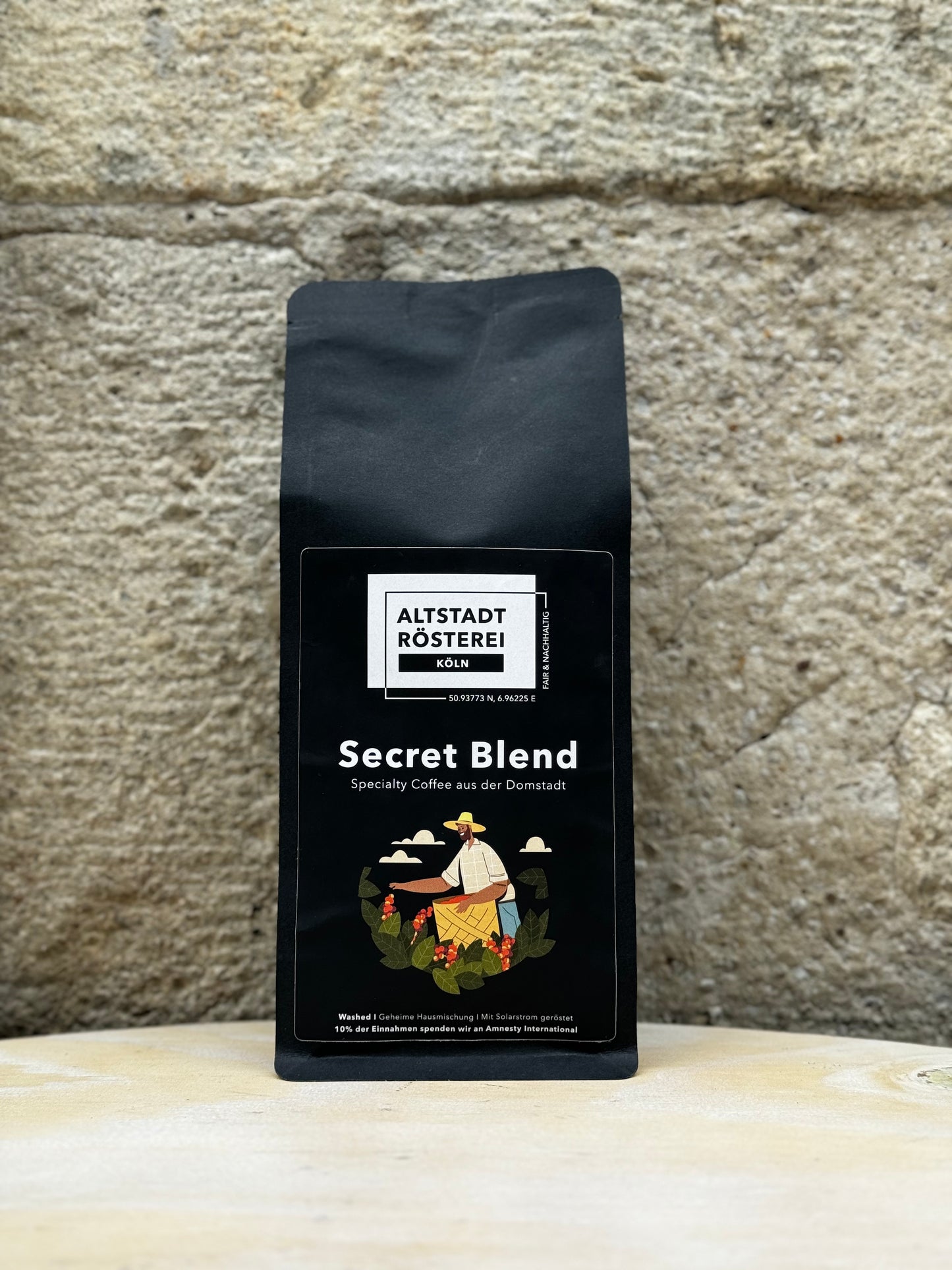 Eine Packung Altstadt Rösterei Köln Secret Blend Kaffee vor einer Steinwand. Auf der Verpackung ist eine Illustration eines Kaffeebauern, der Kaffeekirschen pflückt, sowie die Beschreibung "Specialty Coffee aus der Domstadt" zu sehen.