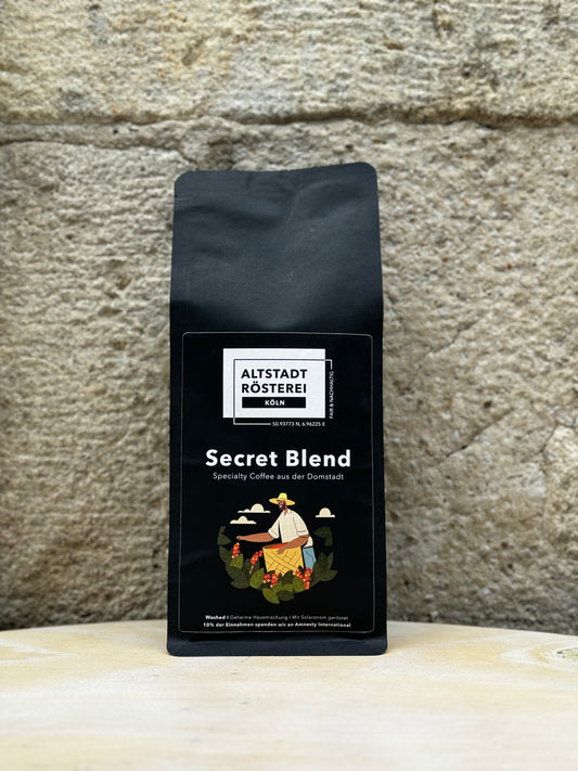 Eine Packung Altstadt Rösterei Köln Secret Blend Kaffee vor einer Steinwand. Auf der Verpackung ist eine Illustration eines Kaffeebauern, der Kaffeekirschen pflückt, sowie die Beschreibung "Specialty Coffee aus der Domstadt" zu sehen.