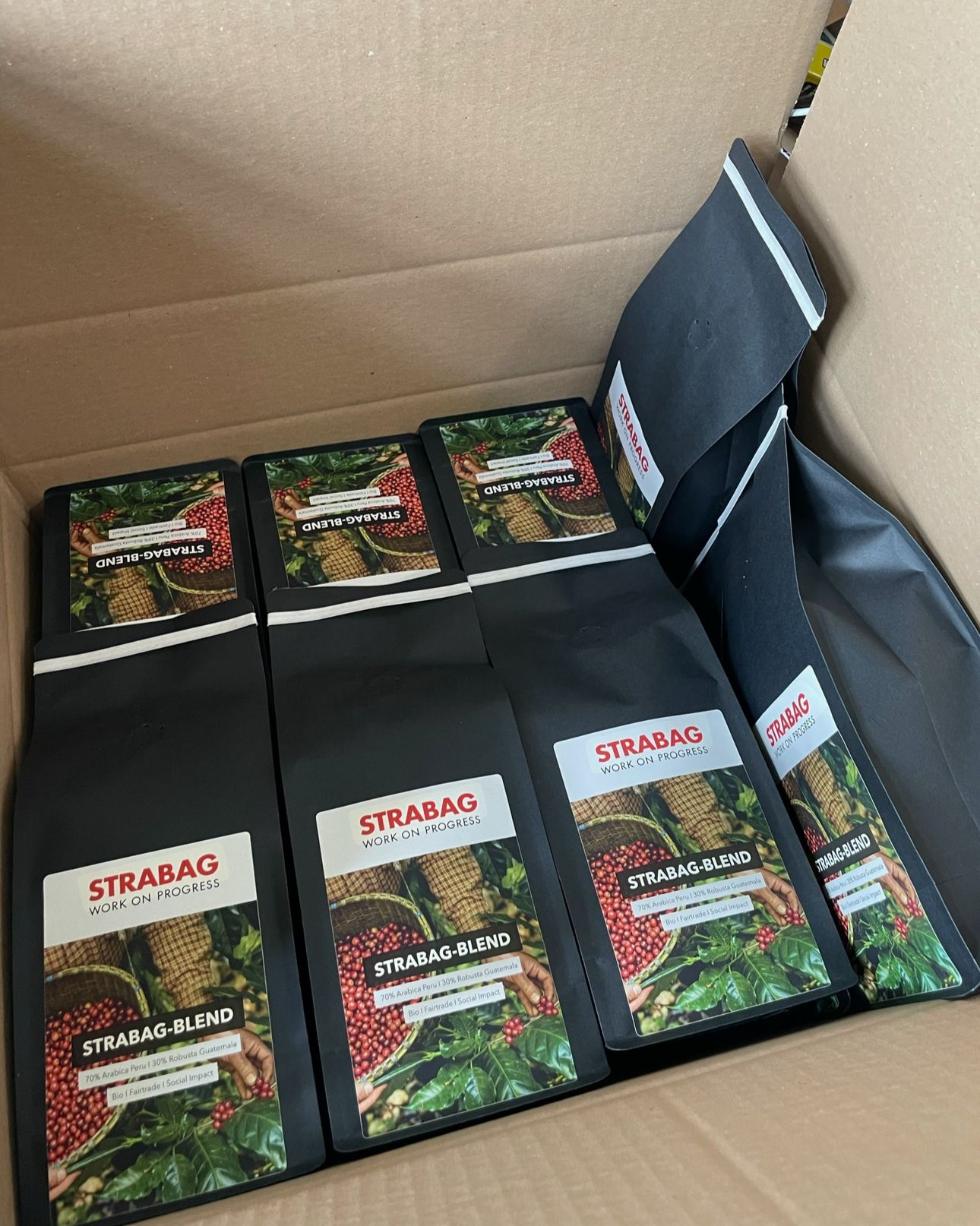 Ein Karton mit mehreren Packungen STRABAG-BLEND Kaffee. Die Kaffeepackungen sind schwarz und haben ein Etikett mit dem STRABAG-Logo sowie einem Bild von Kaffeekirschen und einem Korb. Auf dem Etikett steht "70% Arabica Peru / 30% Robusta Guatemala", "Bio", "Fairtrade" und "Social Impact".