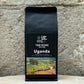 Ein Bild des Produkts Uganda 2100% Robusta von True Beans auf einem Holztisch vor einer steinernen Säule.