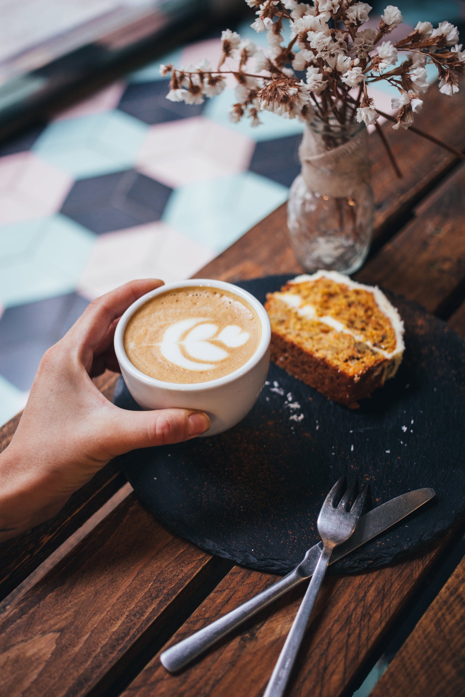 Eine Hand hält eine Tasse Latte mit Latte Art auf einem Tisch, daneben liegt ein Stück Kuchen auf einem Teller, mit Besteck und einer Vase mit getrockneten Blumen im Hintergrund.