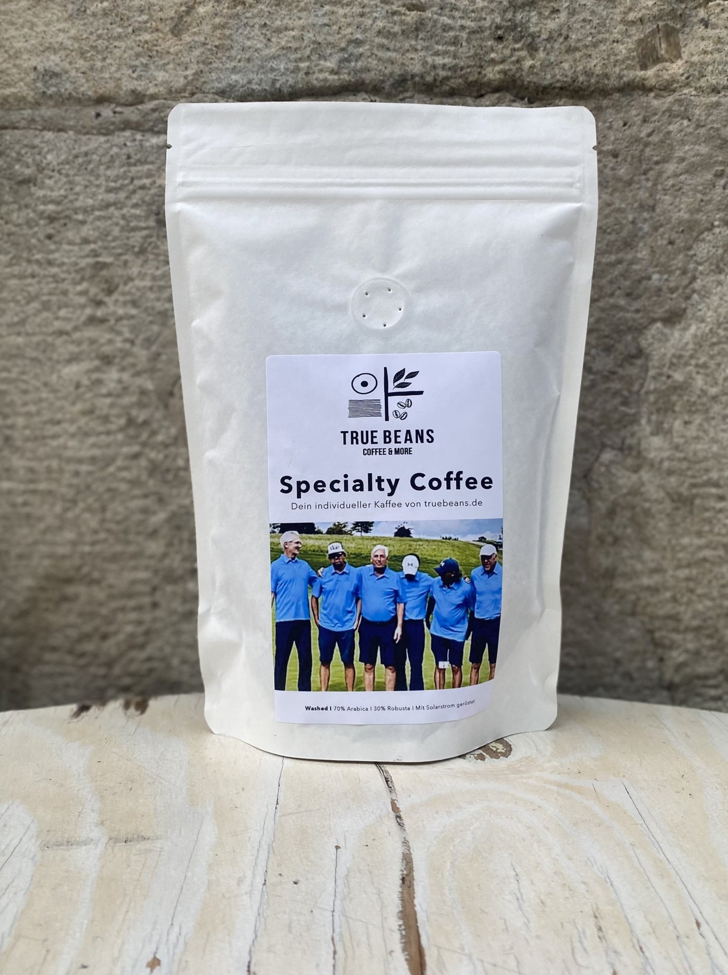 Das Bild zeigt eine weiße Packung Specialty Coffee von True Beans. Auf der Packung ist ein Etikett mit dem True Beans-Logo und einem Foto einer Gruppe von Menschen in blauen Hemden zu sehen. Die Packung steht auf einer hellen Holzoberfläche vor einer grob strukturierten Steinwand.