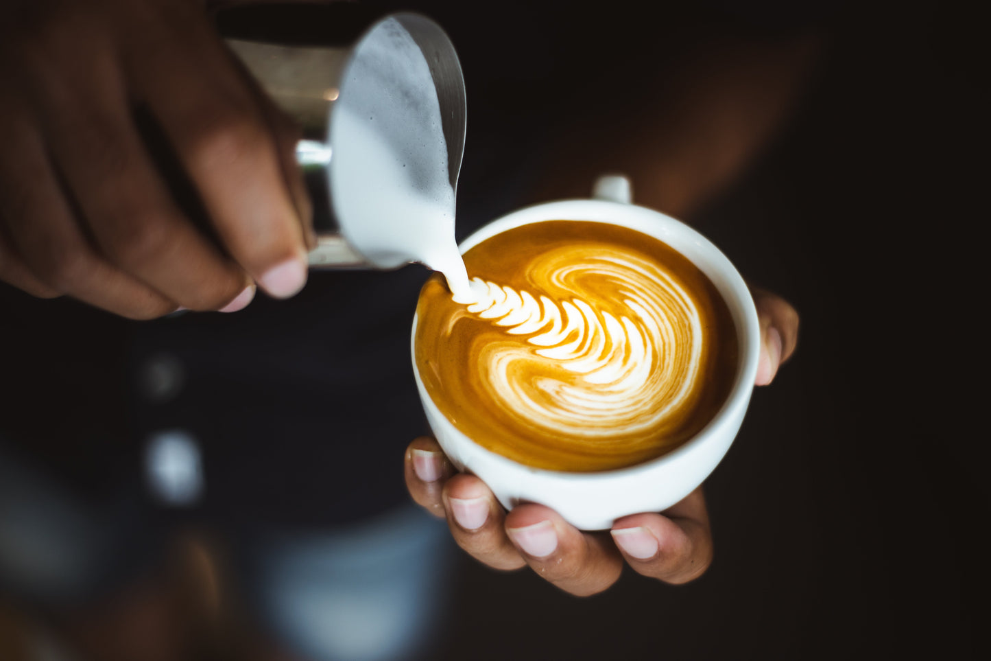 Eine Person gießt geschäumte Milch aus einem silbernen Kännchen in eine Tasse mit Kaffee, um Latte Art zu erstellen. Die Latte Art zeigt ein elegantes, federförmiges Muster auf der Oberfläche des Kaffees.