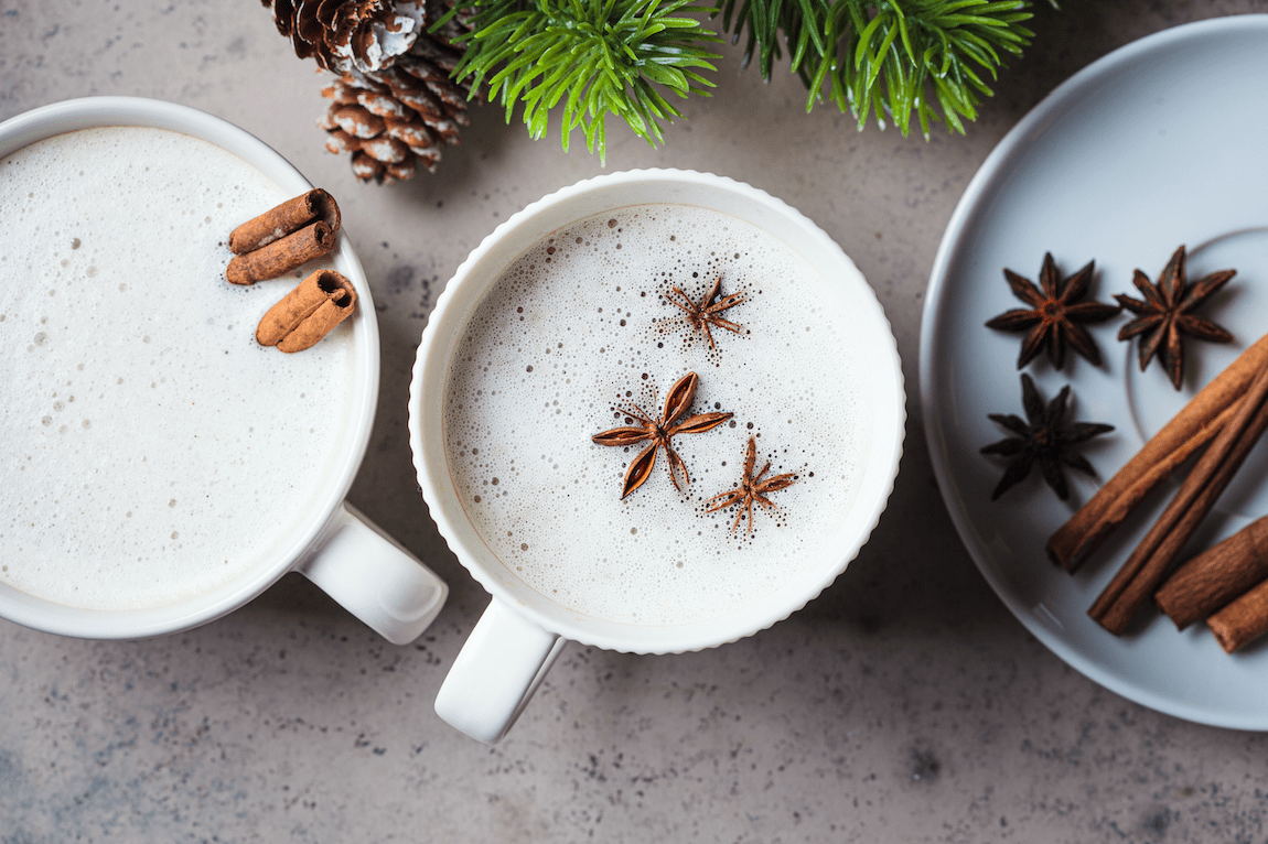 Zwei weiße Tassen gefüllt mit einem cremigen Getränk, dekoriert mit Sternanis und Zimtstangen, stehen auf einer grauen Oberfläche. Im Hintergrund sind Tannenzweige und eine Tannenzapfe zu sehen. Auf einem weißen Teller daneben liegen weitere Zimtstangen und Sternanis.